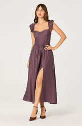 A woman is wearing a long, purple dress with textured fabric. The dress features a fitted bodice with sweetheart neckline and ruffled shoulder straps. The skirt is flowing with a high slit on the left side. She is wearing brown high-heeled shoes.
